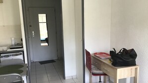Quarto pequeno e limpo, com secretária, cadeira e cama. Há uma porta que dá para outro espaço e uma janela que ilumina naturalmente.
