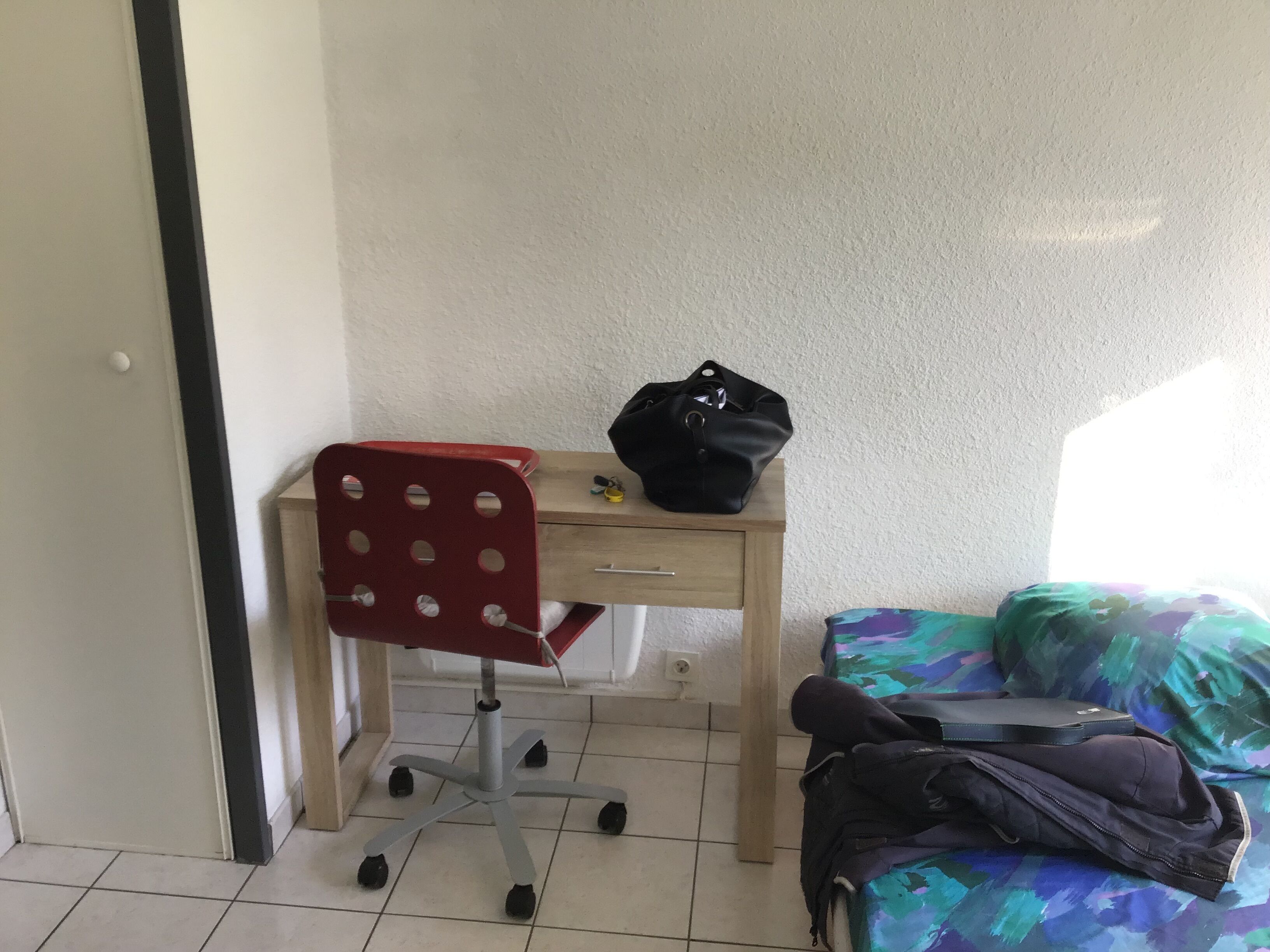 A small room with a desk, a red office chair, a black bag on the desk, and a bed with a colorful blanket.