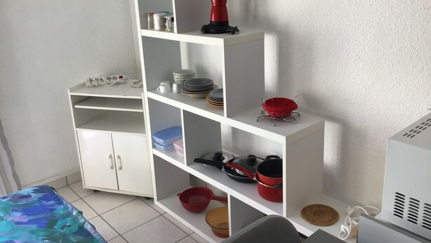 Uma cozinha com armários brancos, bancada branca e parede branca. Há uma chaleira vermelha sobre a bancada.