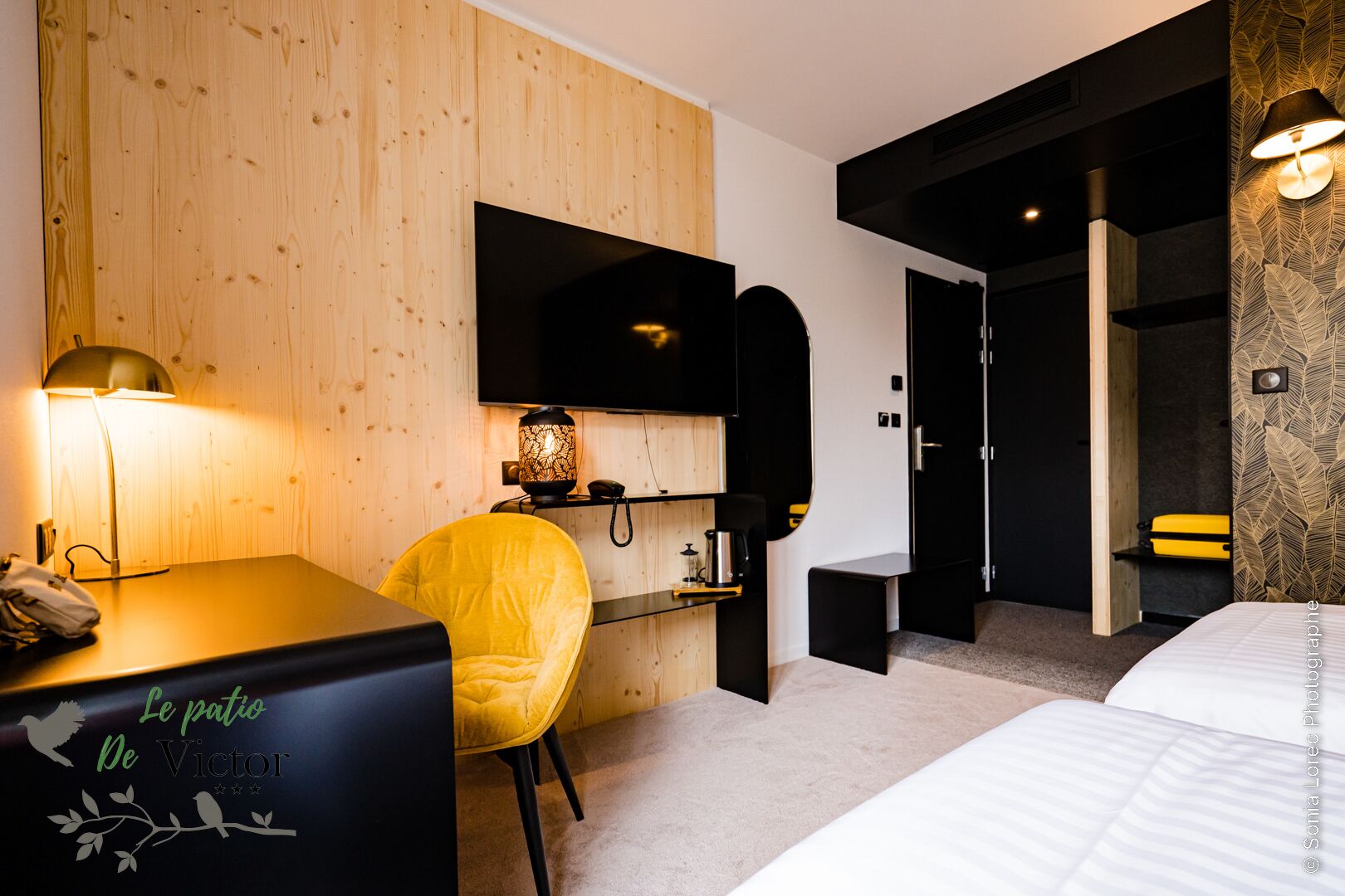 A modern hotel room with a wooden wall, a desk with a lamp, a yellow chair, a bed, and a TV mounted on the wall.
