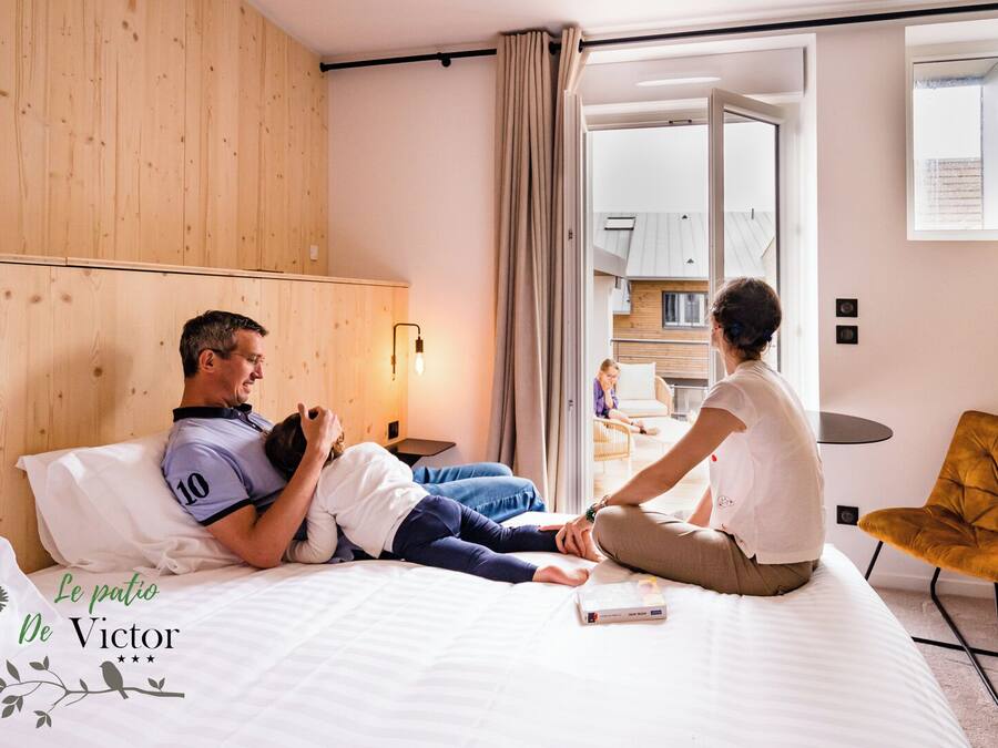 A hotel room with a wooden headboard, a bed with white bedding, a bedside table with a lamp, and a view of the kitchen through an open door.