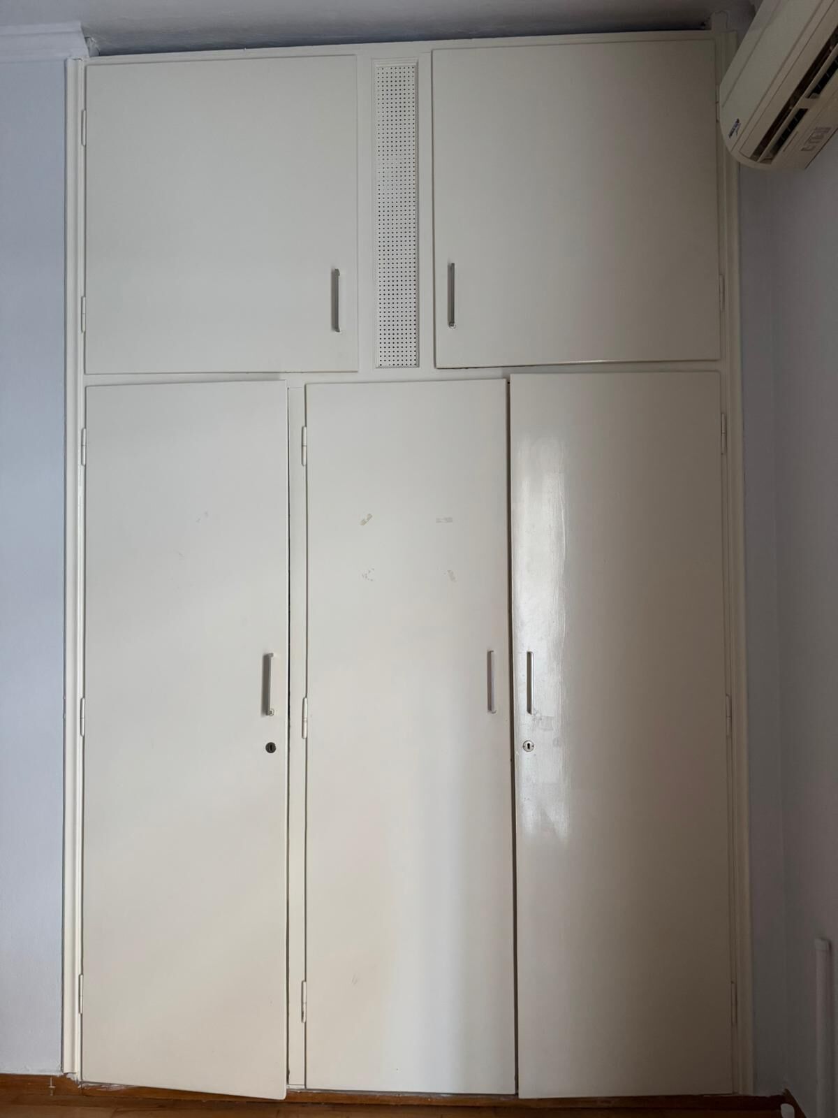 A white wardrobe with two doors and two drawers, a ventilation panel, and an air conditioning unit on the wall.
