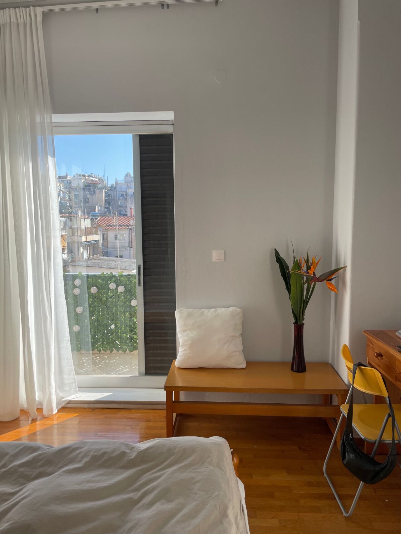 Une chambre à coucher avec un lit, un banc, une chaise, une petite table, une plante et une vue sur la ville par la fenêtre.