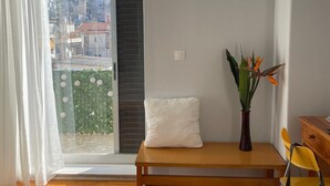 Une chambre à coucher avec un lit, un banc, une chaise, une petite table, une plante et une vue sur la ville par la fenêtre.