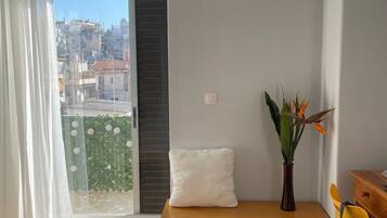 Ein Schlafzimmer mit einem Bett, einer Bank, einem Sessel, einem kleinen Tisch, einer Pflanze und Blick auf die Stadt durch ein Fenster.