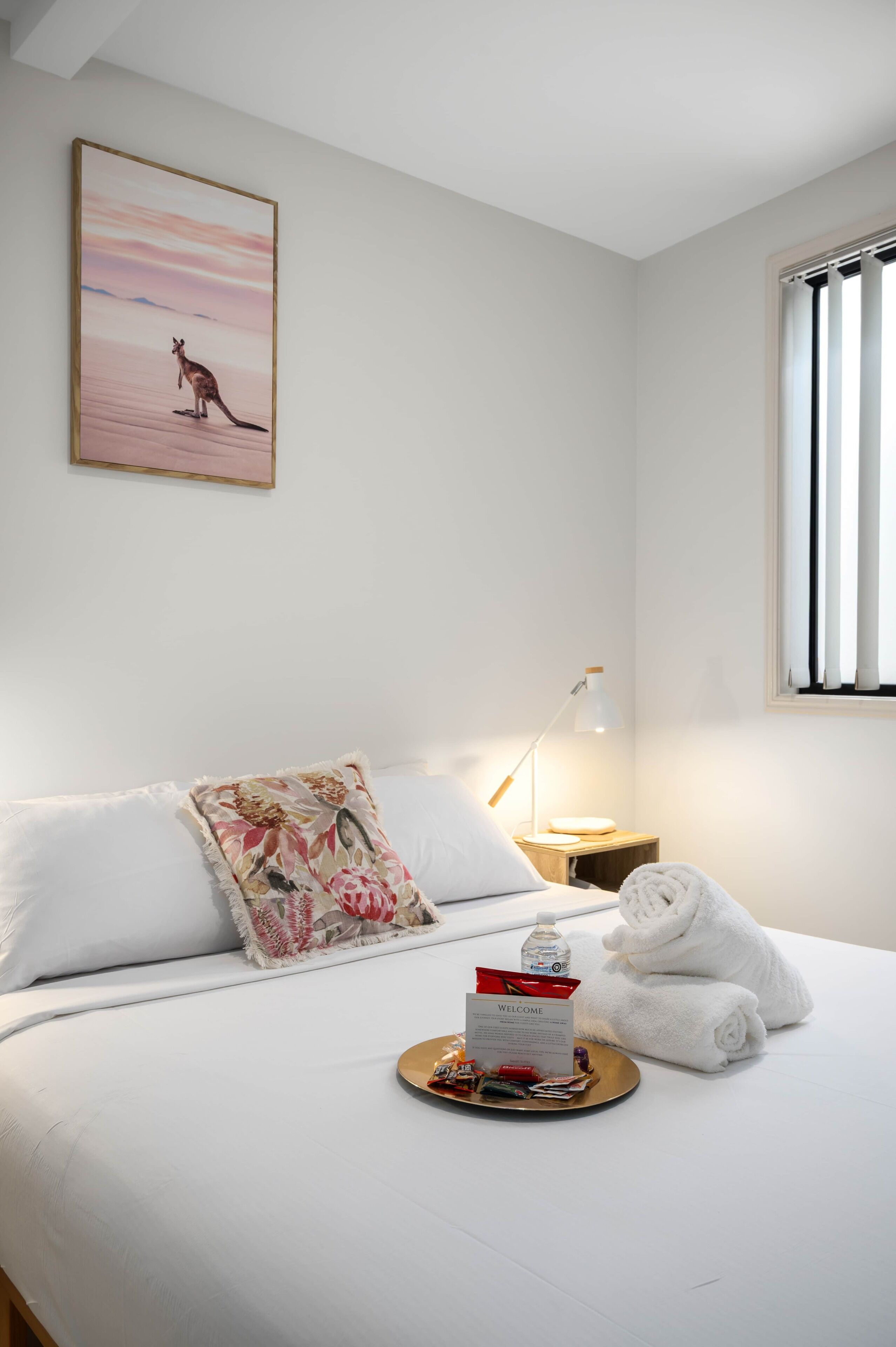 Cama bem arrumada com lençóis brancos, um travesseiro floral, um bandeja com um cartão de boas-vindas e toalhas.