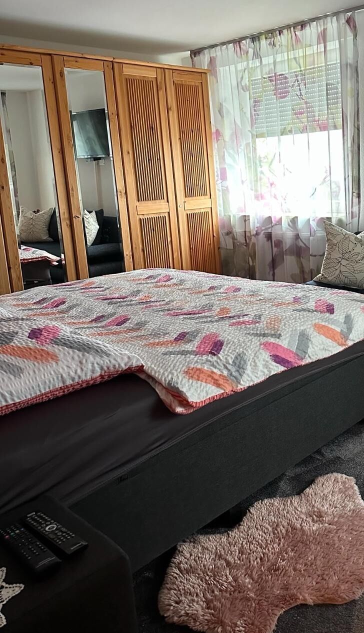 A bed with a patterned quilt, a wooden wardrobe, a mirror, and a window with floral curtains.