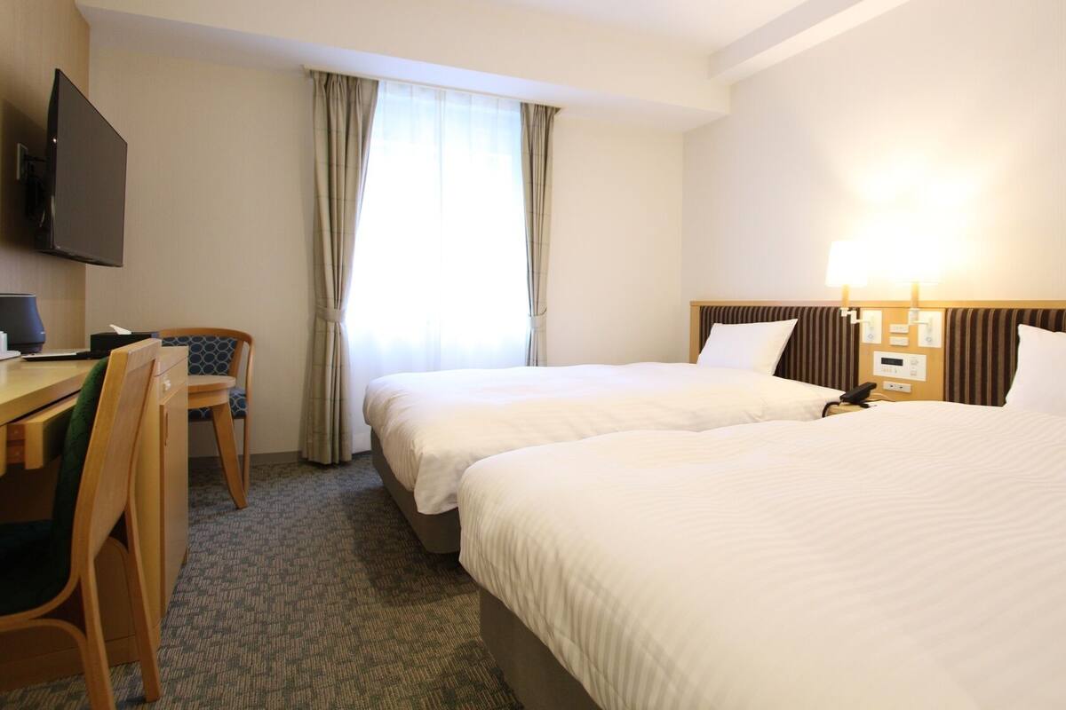 Een hotelkamer met twee bedden, een bureau met stoel, een televisie en een raam met gordijnen.