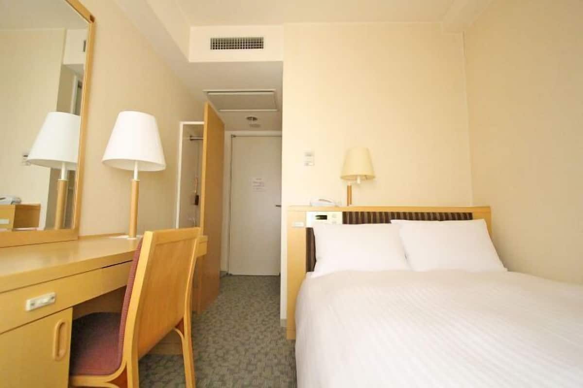 A hotel room with a bed, a desk, a chair, a mirror, and a lamp.