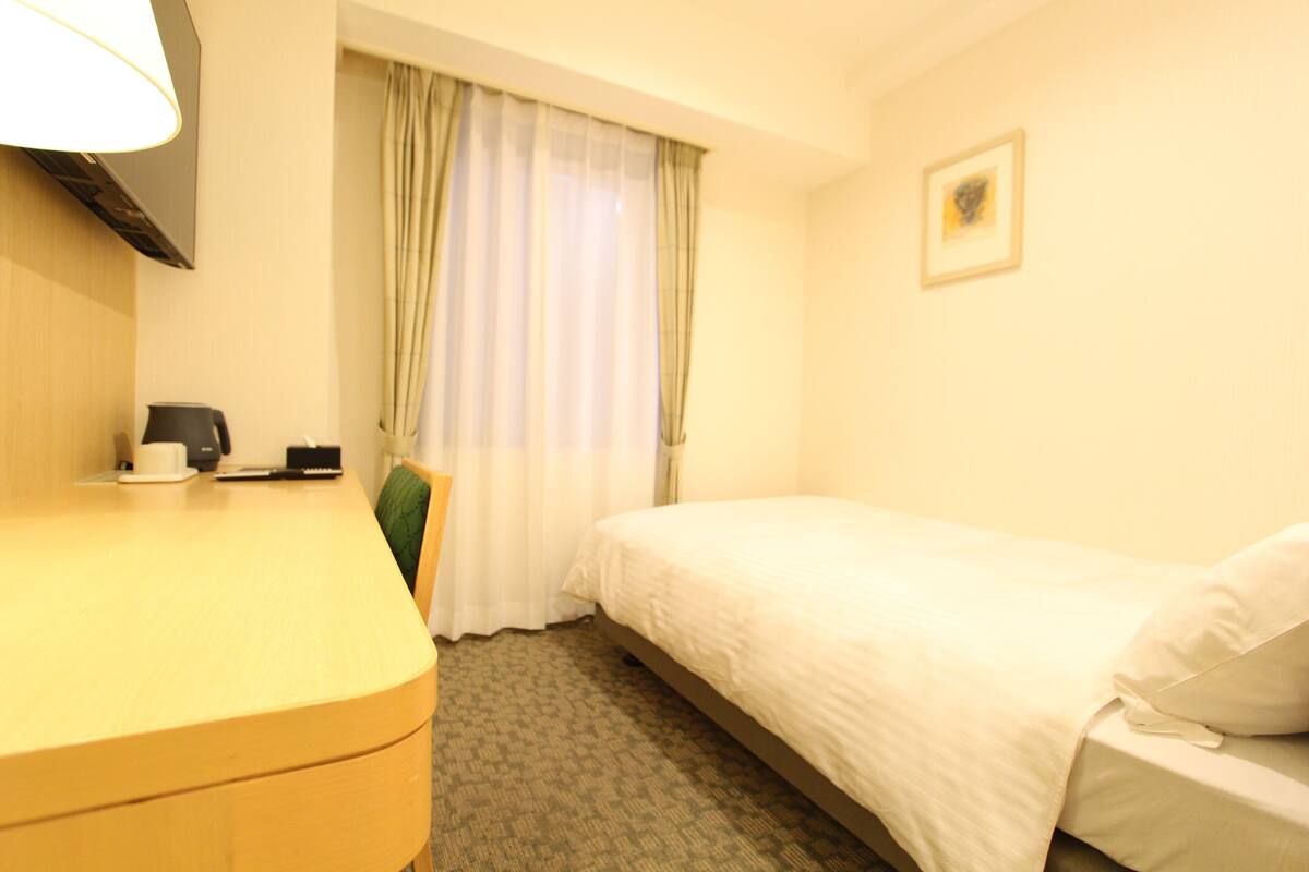 Hotelkamer met een bed, een bureau, een stoel, een lamp, een foto aan de muur en een raam met gordijnen.