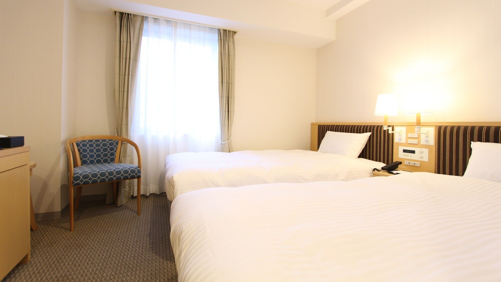Een hotelkamer met twee bedden, een stoel, een bureau, een lamp en een raam met gordijnen.