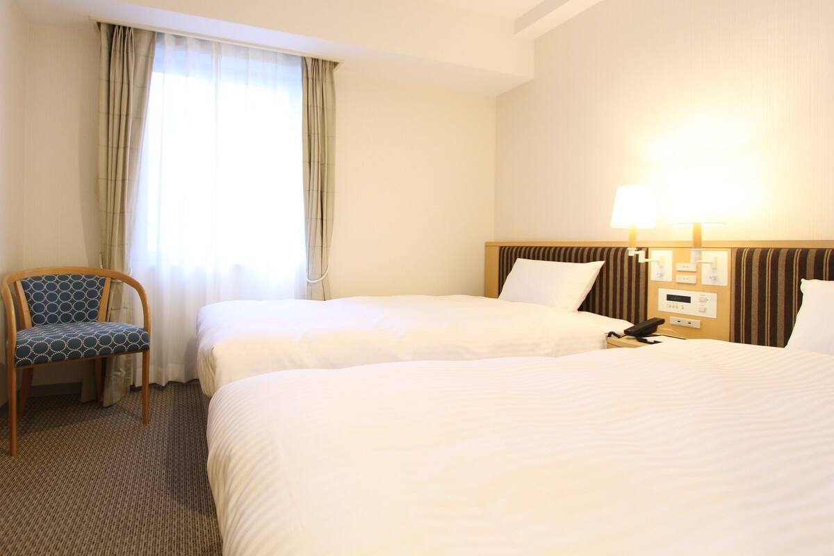 Een hotelkamer met twee bedden, een stoel, een bureau, een lamp en een raam met gordijnen.