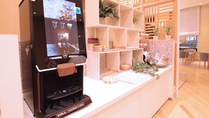 A coffee machine with a touchscreen interface, a white counter with shelves containing various items, and a wooden partition with plants. - KOKO HOTEL Sendai Station South (Sendai)
