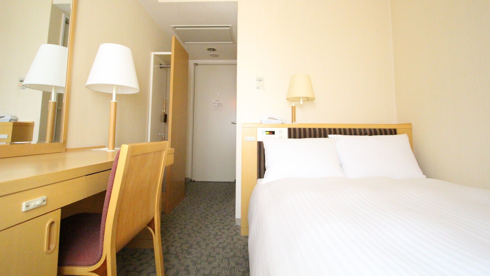 Een hotelkamer met een bed, een bureau met stoel, een spiegel en een deur naar een andere kamer.