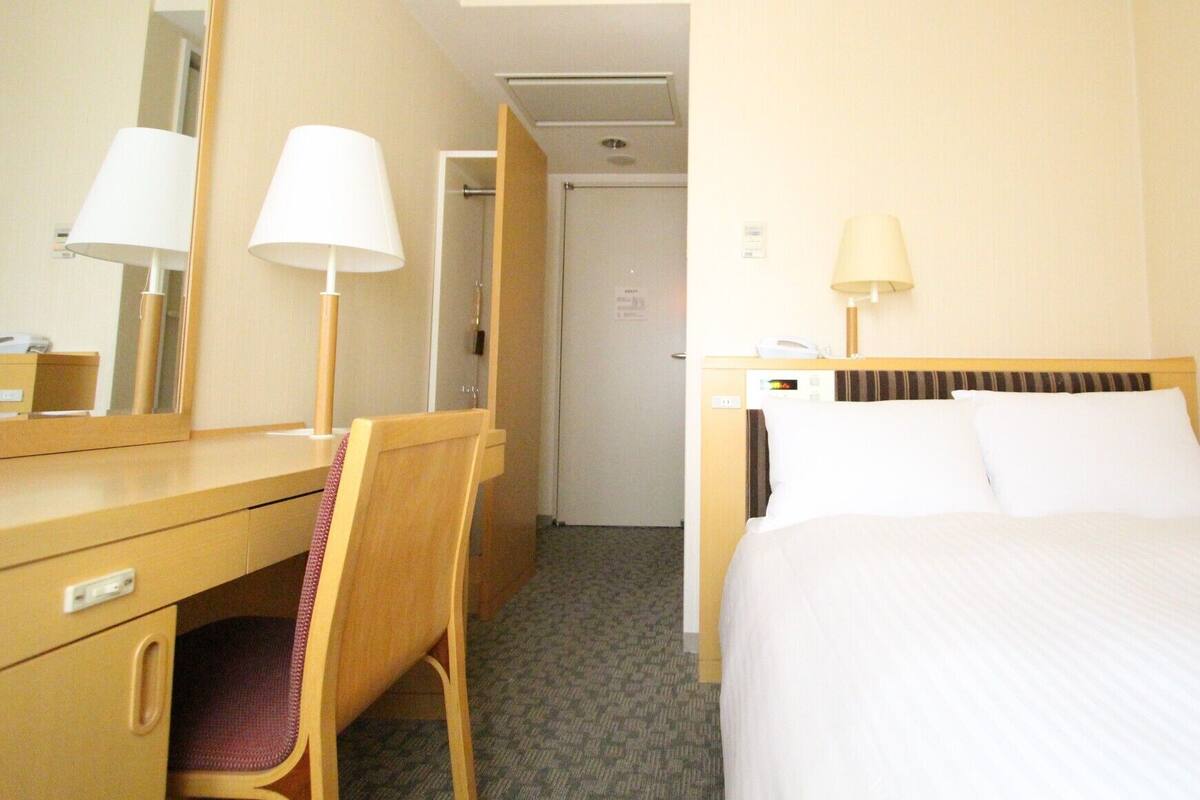 Een hotelkamer met een bed, een bureau met stoel, een spiegel en een deur naar een andere kamer.