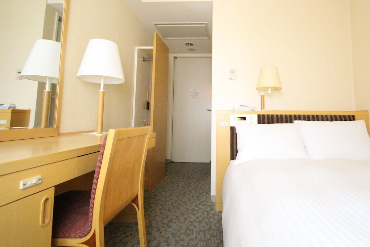 A hotel room with a bed, a desk with a chair, a mirror, and a door leading to another room.