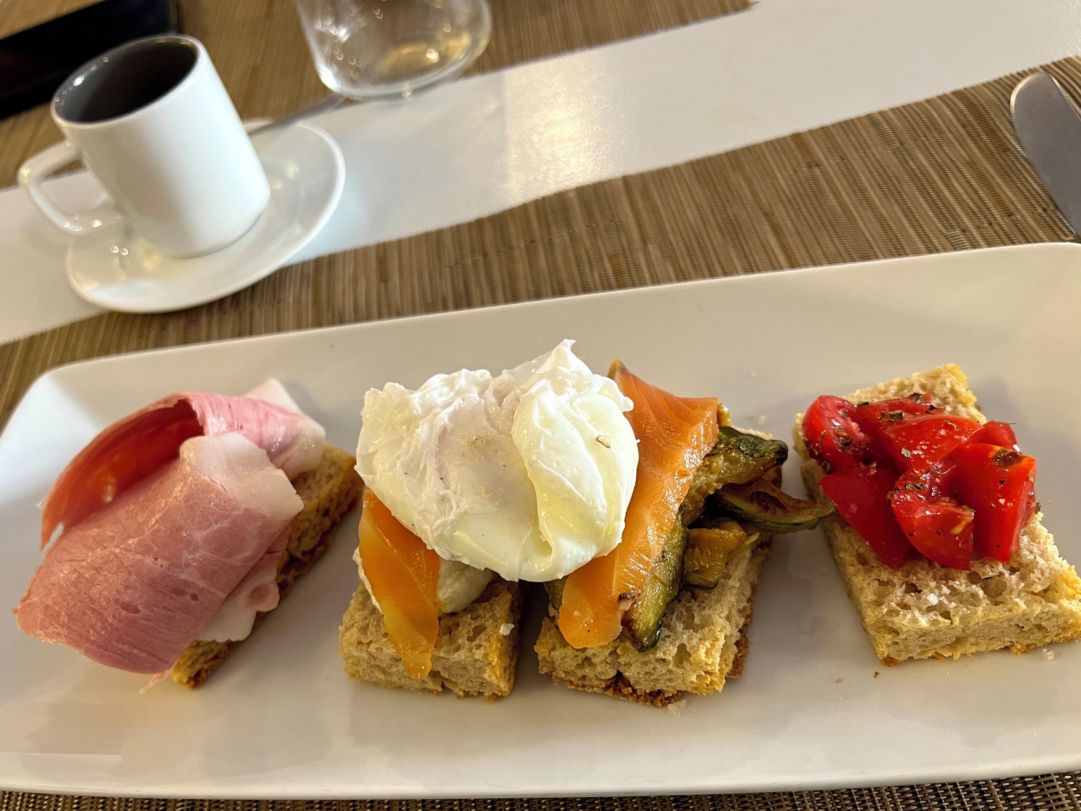 A white rectangular plate with three different food items: a slice of bread with smoked salmon and a poached egg, a slice of bread with smoked salmon and grilled vegetables, and a slice of bread with tomato slices.