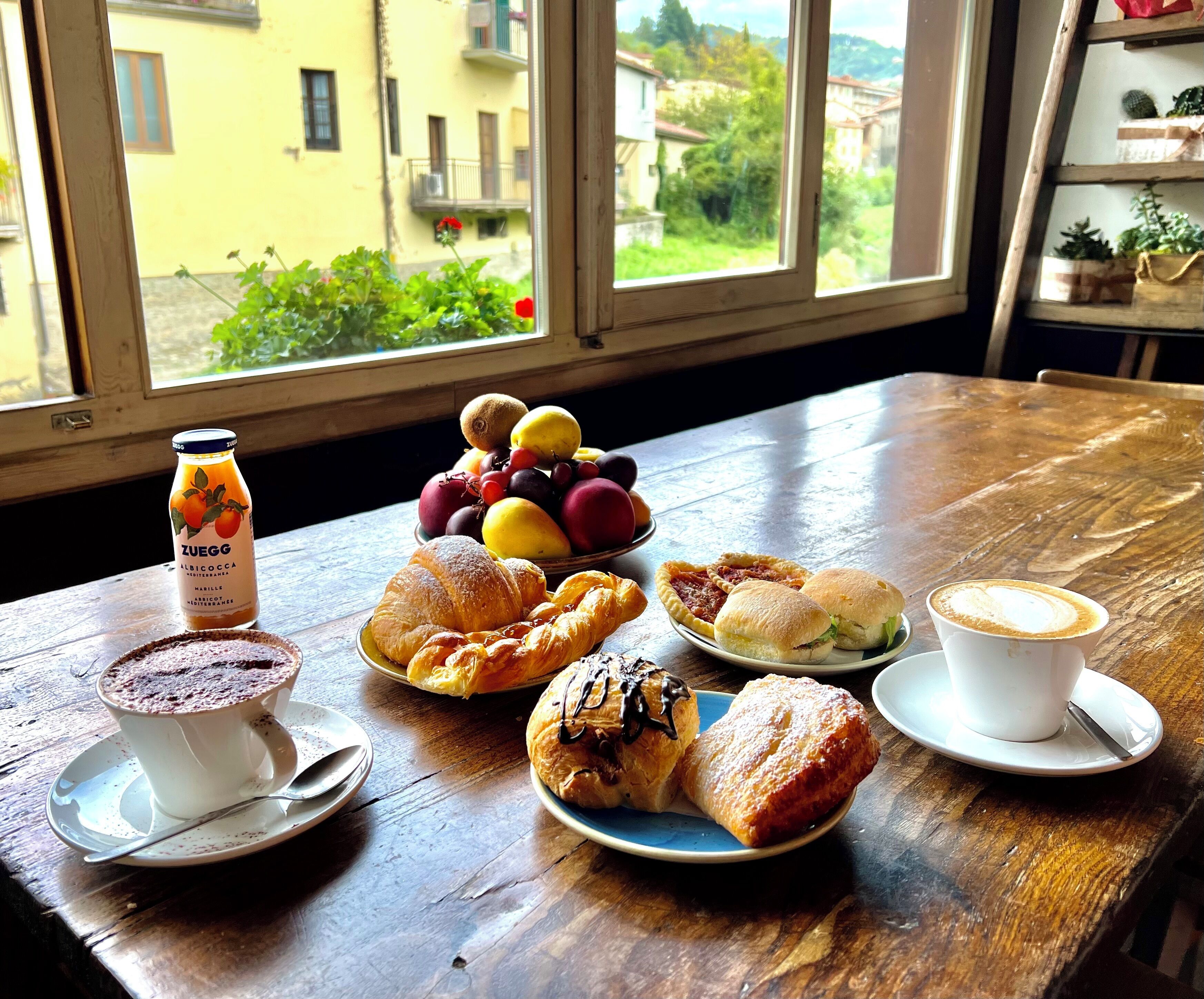 Una mesa de desayuno con cruasanes, bollería, un bol de fruta, una botella de jugo y una taza de café.