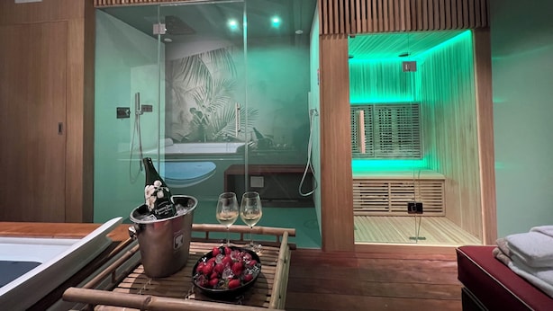 A modern spa room with a glass-enclosed shower, a wooden bench with a tray of strawberries and champagne, and a green-lit relaxation area. - Nobilia Dimore - Suite & Spa (Pratovecchio Stia)