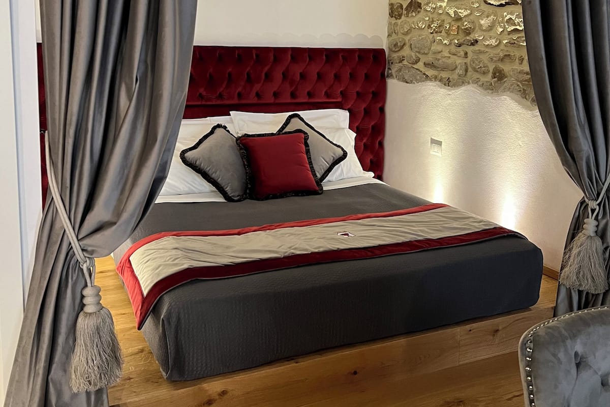 Quarto com cabeceira estofada vermelha, cortinas cinzentas e parede de pedra.