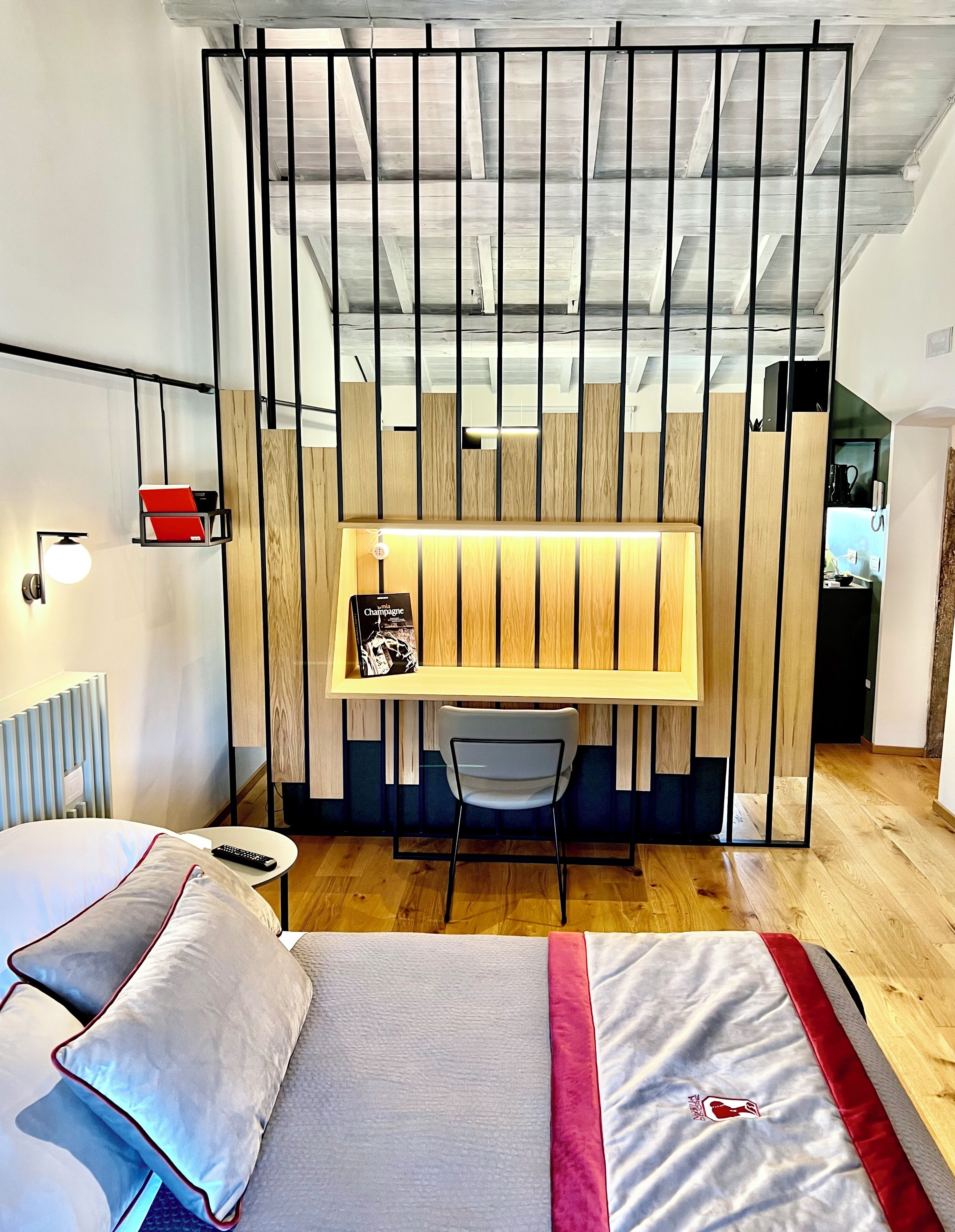 A compact hotel room with a bed, a desk, and a chair. The room features a wooden partition with vertical slats and a small shelf with a book.