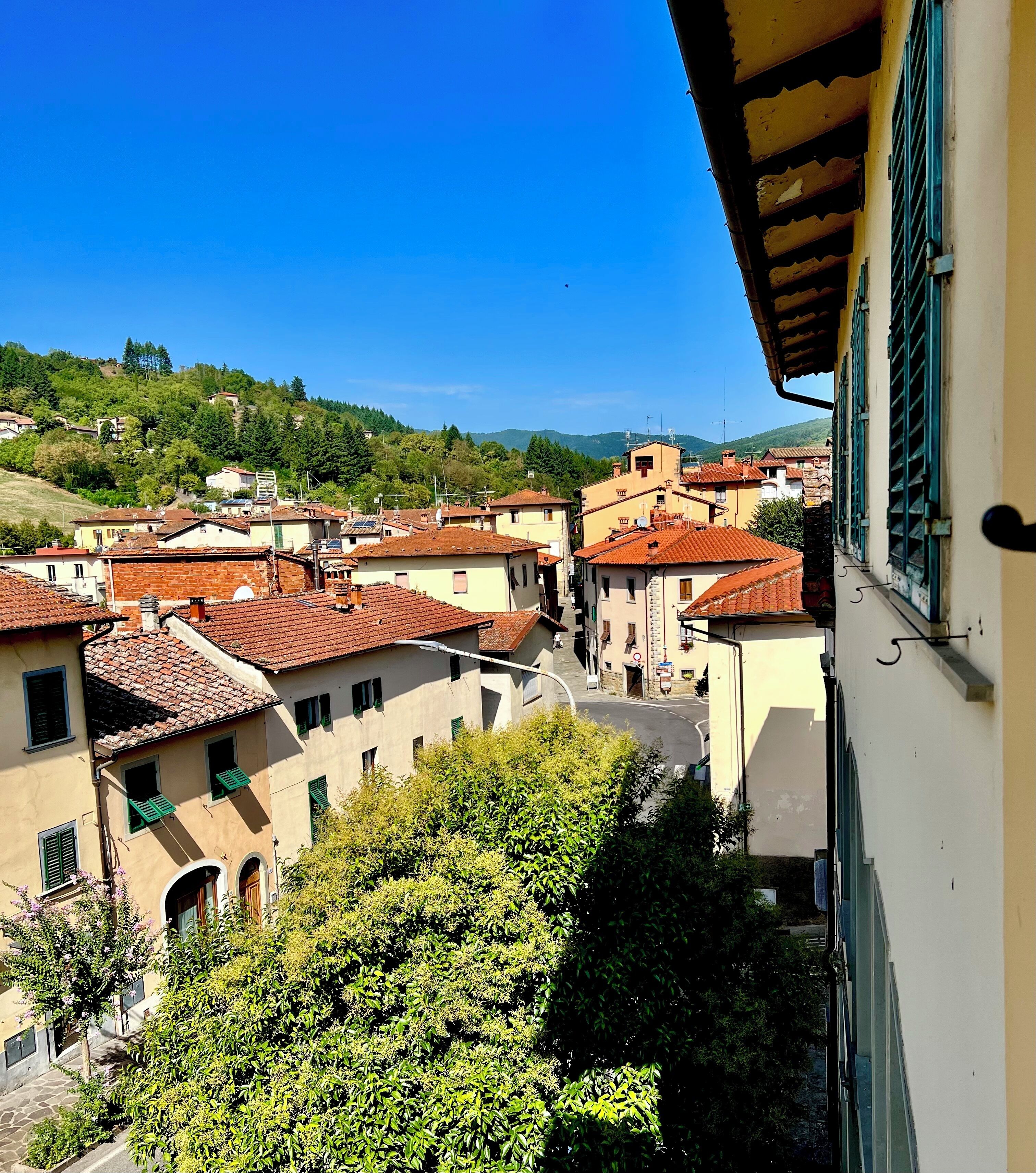 Une vue sur une ville perchée sur une colline, avec des bâtiments aux toits de tuiles rouges, un ciel bleu dégagé et une végétation luxuriante.