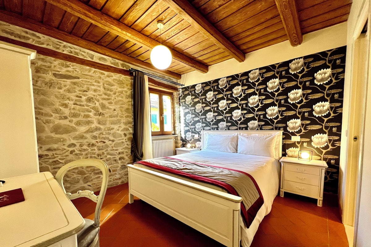 Quarto com parede de pedra, teto de madeira, cama com roupa de cama branca, mesa de cabeceira e uma cadeira.
