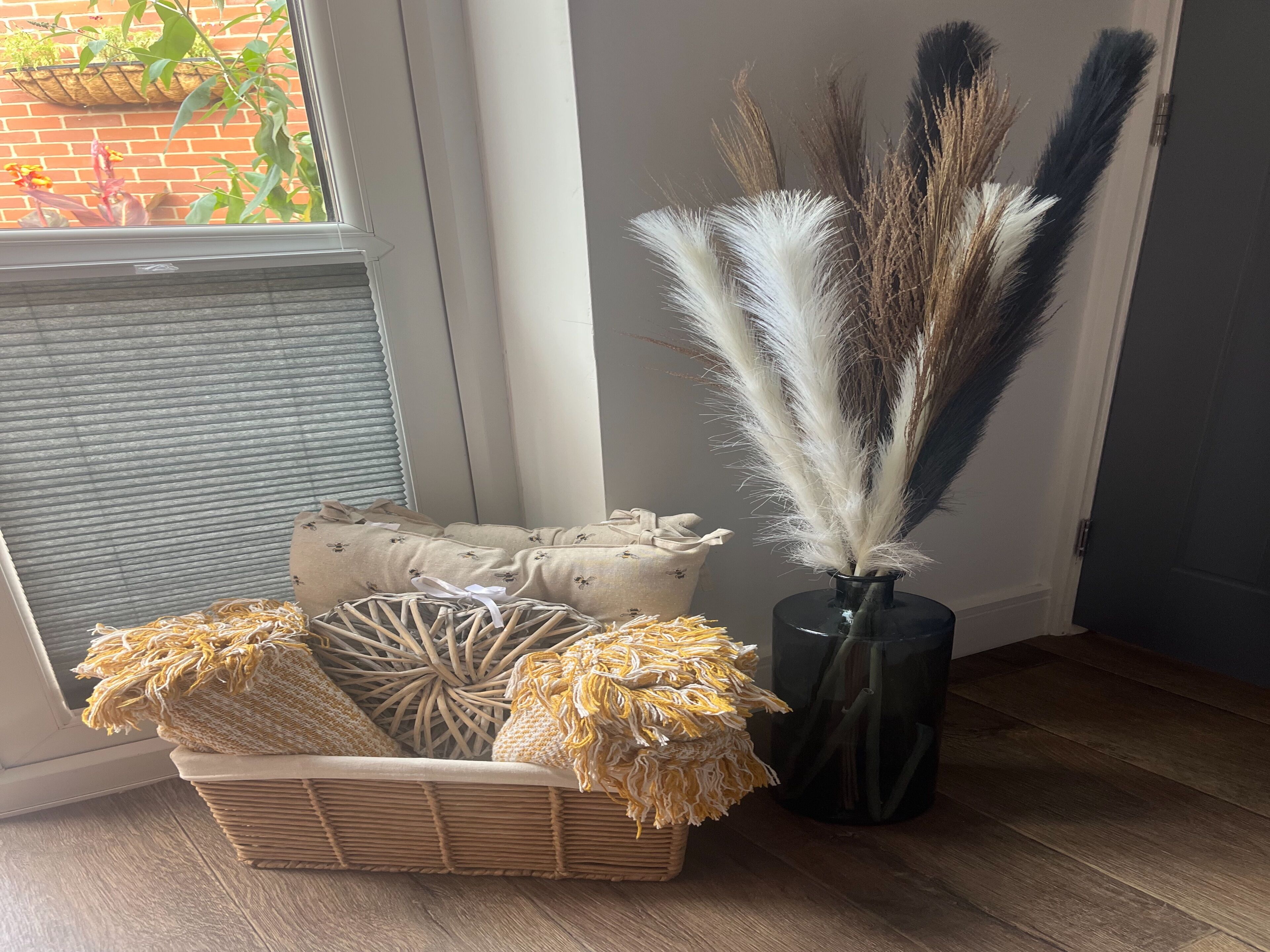 A corner of a room with a wicker basket containing decorative items, a vase with tall feathers, and a window with a view of a brick wall and greenery outside.