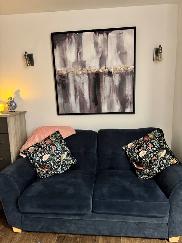 A dark blue sofa with floral-patterned cushions, a large abstract painting on the wall, and a small table with a vase and candle. - Stunning 1-bed Private Apartment in Norwich (Norwich)