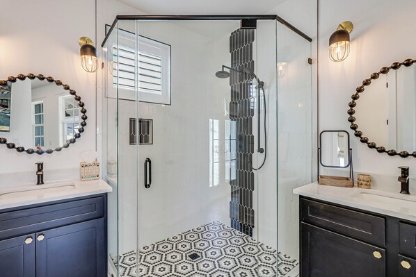 A modern bathroom with a glass shower enclosure, a white tiled floor, and two sinks with dark cabinets.