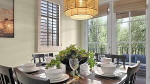 Una mesa de comedor dispuesta con platos, fuentes y cubiertos blancos, una copa y un centro de mesa con follaje verde.