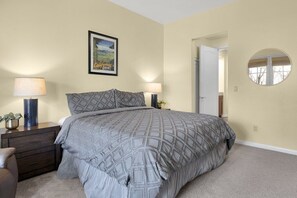 Een slaapkamer met een bed, twee nachtkastjes, een spiegel en een deur naar een andere kamer, plus een lijst met een afbeelding.