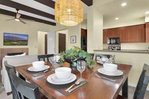 A dining area with a wooden table set for four, surrounded by chairs, with a large plant centerpiece and a ceiling fan above. - 3BR Legacy Villas condo with pool, patio, Wi-Fi, and mountain charm (La Quinta)