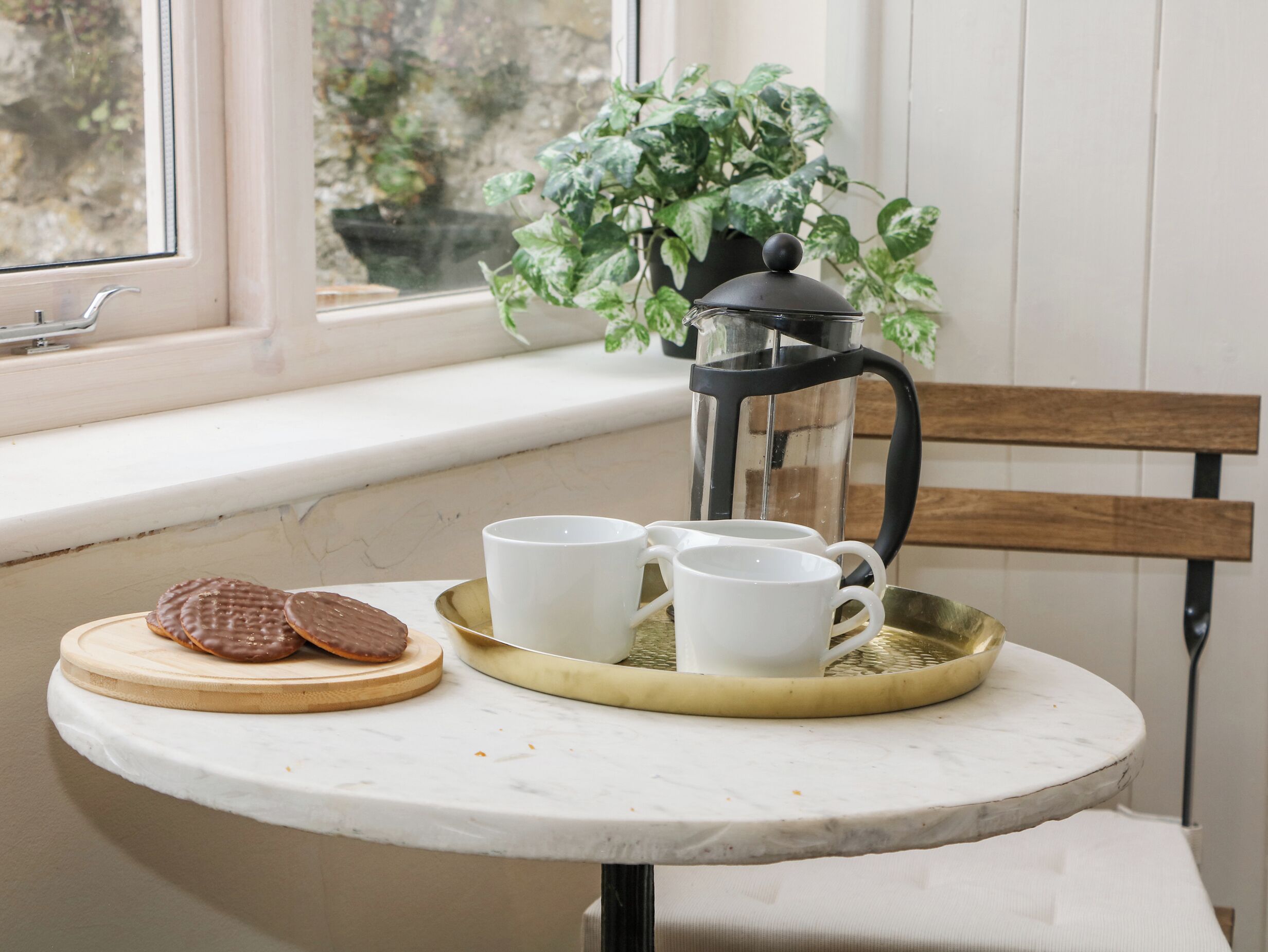 A small round table with a white marble top, a tray with two white cups and a black coffee pot, and a wooden cutting board with two chocolate-covered cookies.