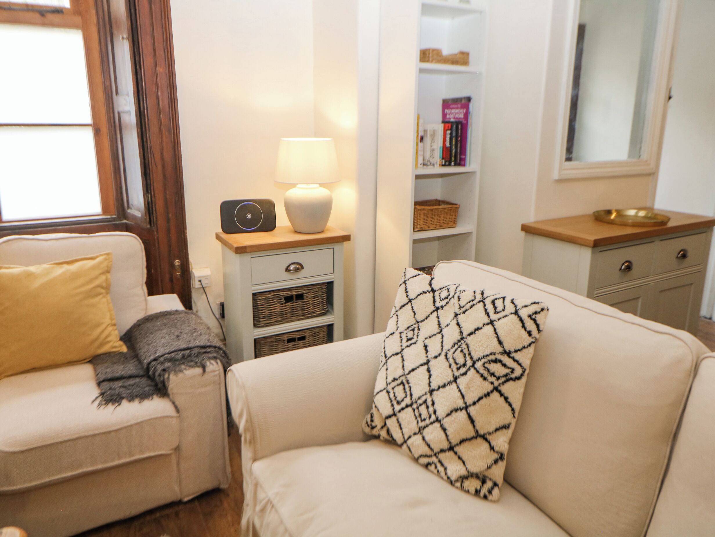 Un salon avec un canapé beige, un fauteuil assorti, une table d’appoint avec une lampe, une bibliothèque et un petit meuble en bois.