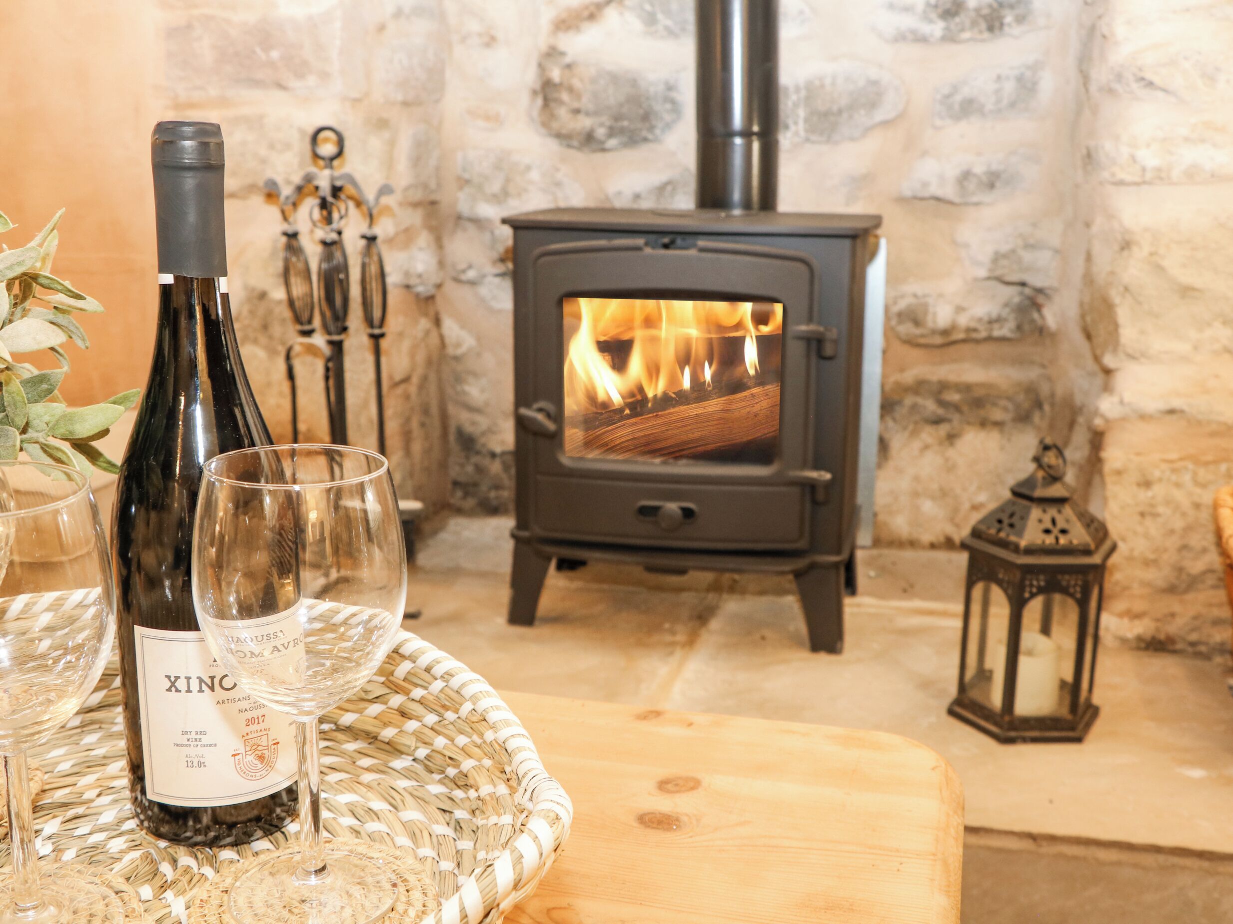 A bottle of XINC wine, two glasses, a woven basket, a wooden cutting board, a stone wall, and a wood-burning stove.