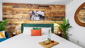 A bedroom with a wooden headboard, a bed with white bedding, a green pillow, and a wooden tray with a bottle and glasses. - Little Havana Retreat, Steps from the Beach - King Size Bed & Gulf Bliss! (Indian Rocks Beach)