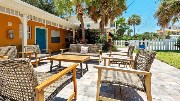 An outdoor seating area with wicker furniture and a wooden table, surrounded by palm trees and a white picket fence. - Little Havana Retreat, Steps from the Beach - King Size Bed & Gulf Bliss! (Indian Rocks Beach)