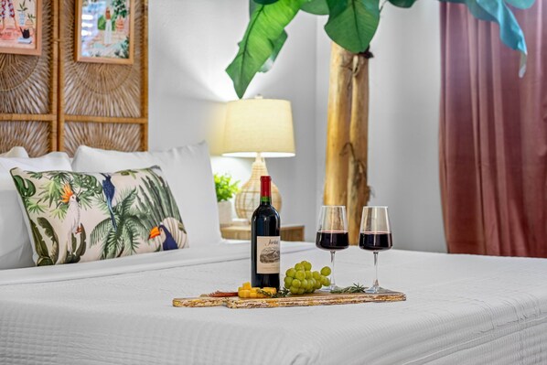 Una cama con una colcha blanca, una botella de vino, dos copas de vino tinto y una bandeja con uvas y queso.