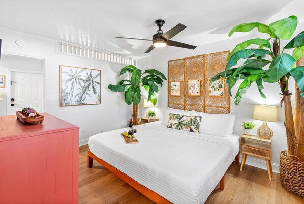 Una habitación de hotel con una cama, un ventilador de techo, una planta grande y una mesita de noche de madera con una lámpara.