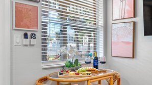 Un coin repas avec une table en osier, un plateau de fruits, du vin et une vue à travers les stores.