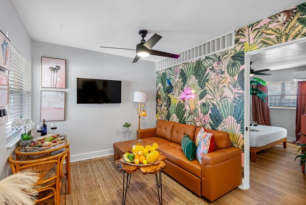A modern living room with a brown leather sofa, a dining table with fruit, a ceiling fan, and a tropical wallpaper.