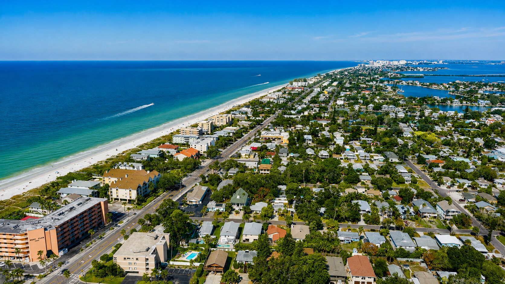 Vue aérienne d’une zone résidentielle côtière, avec une plage, des habitations densément regroupées et une marina.