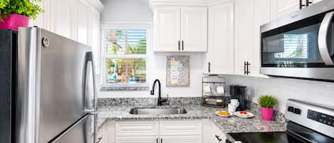 A modern kitchen with white cabinets, stainless steel appliances, a window with blinds, and a coffee station.
