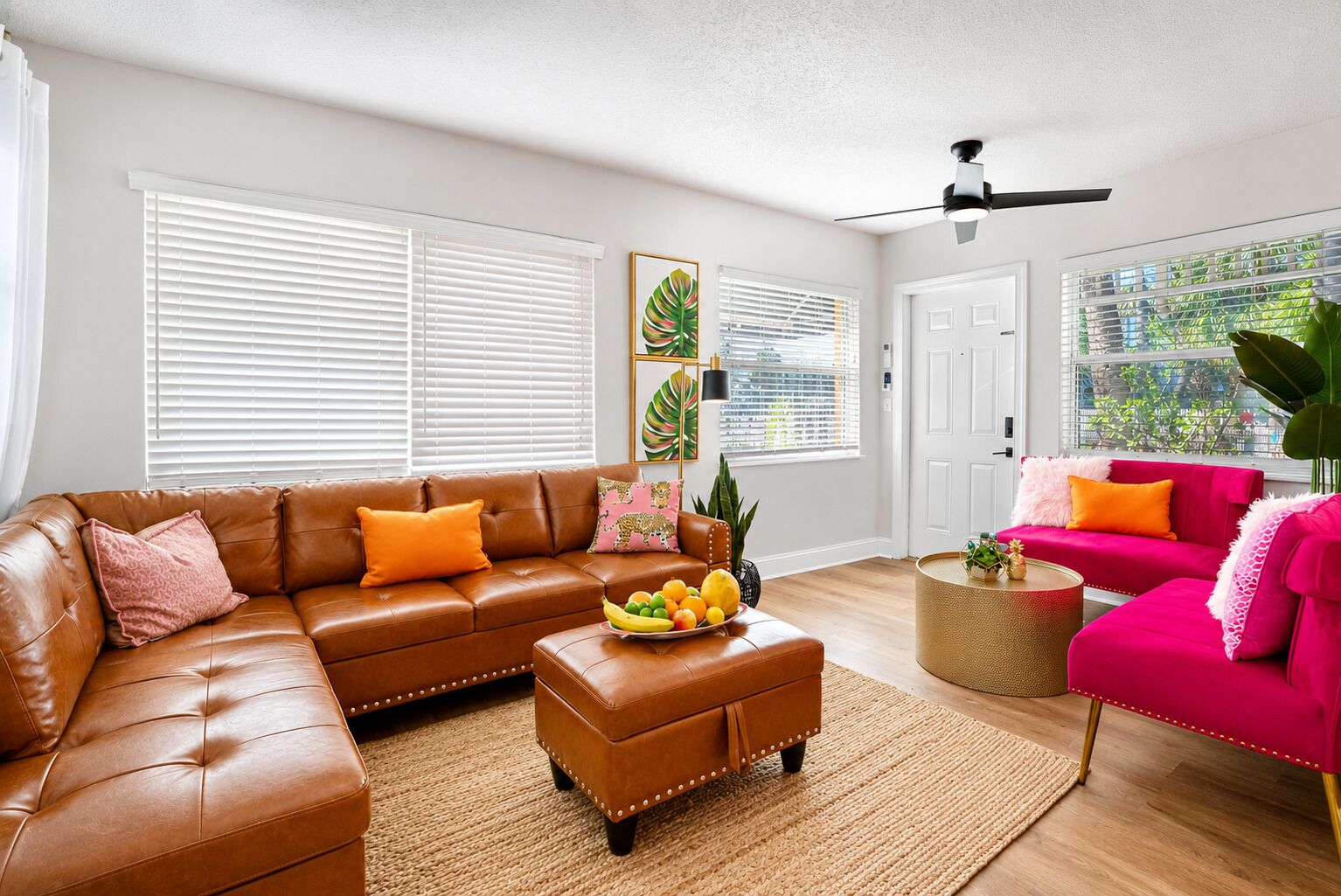 Ein Wohnzimmer mit braunem Ledersofa, einem rosa Sofa, einem Couchtisch mit Obst und einem Deckenventilator.