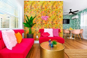 Een moderne woonkamer met levendig tropisch behang, een rode bank, een ronde gouden salontafel en een eethoek met een witte tafel en roze stoelen.
