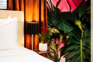 Ein Hotelzimmer mit einem Bett, einem Nachttisch mit Lampe und großen, tropischen Pflanzen.