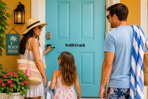 A family approaching a turquoise door labeled 'South Beach' with a beach house sign and a lantern on the wall.