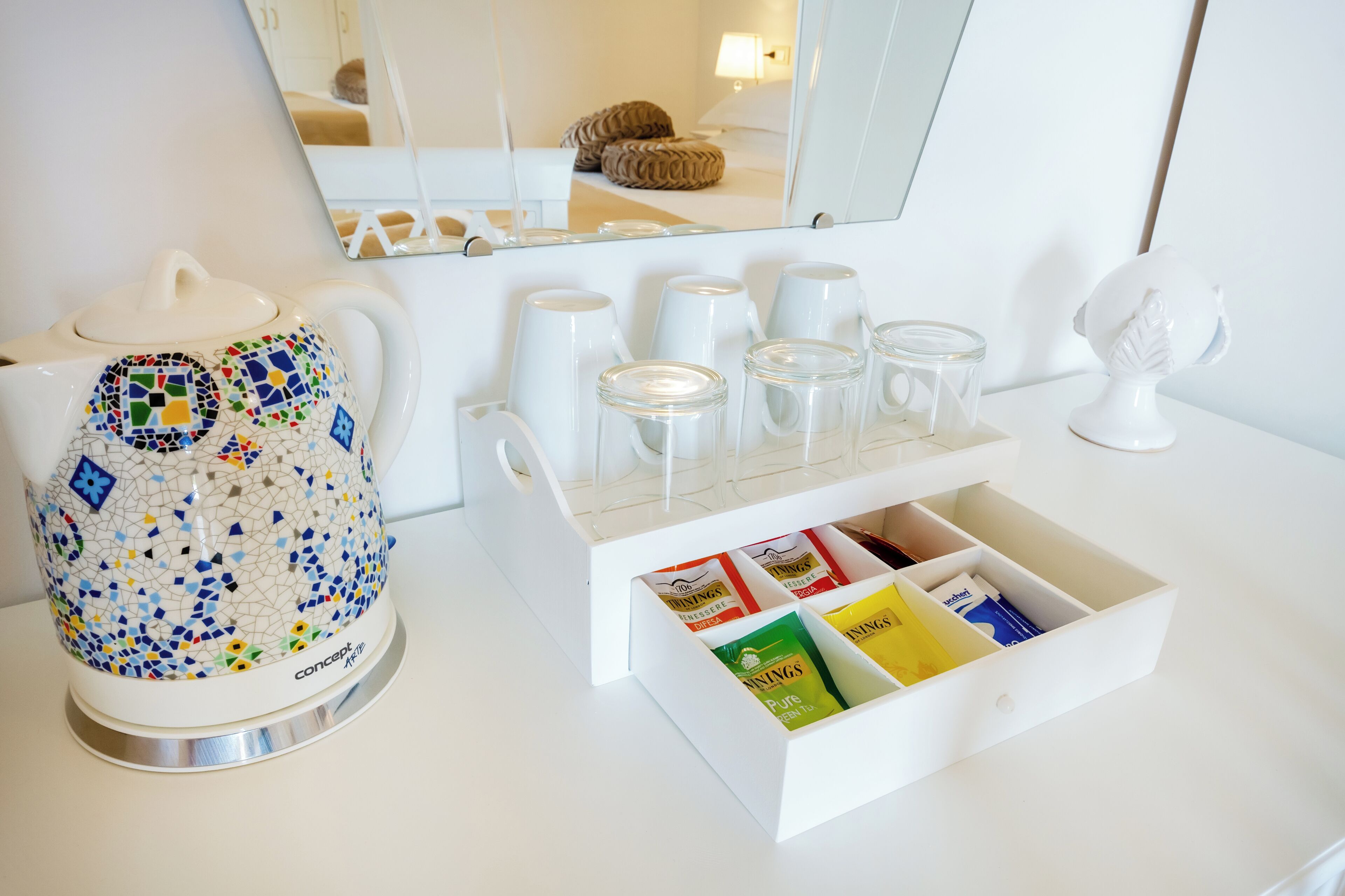 Un plan de travail blanc orné d’une théière à motifs en mosaïque, un ensemble de verres transparents et un tiroir contenant des sachets de thé.