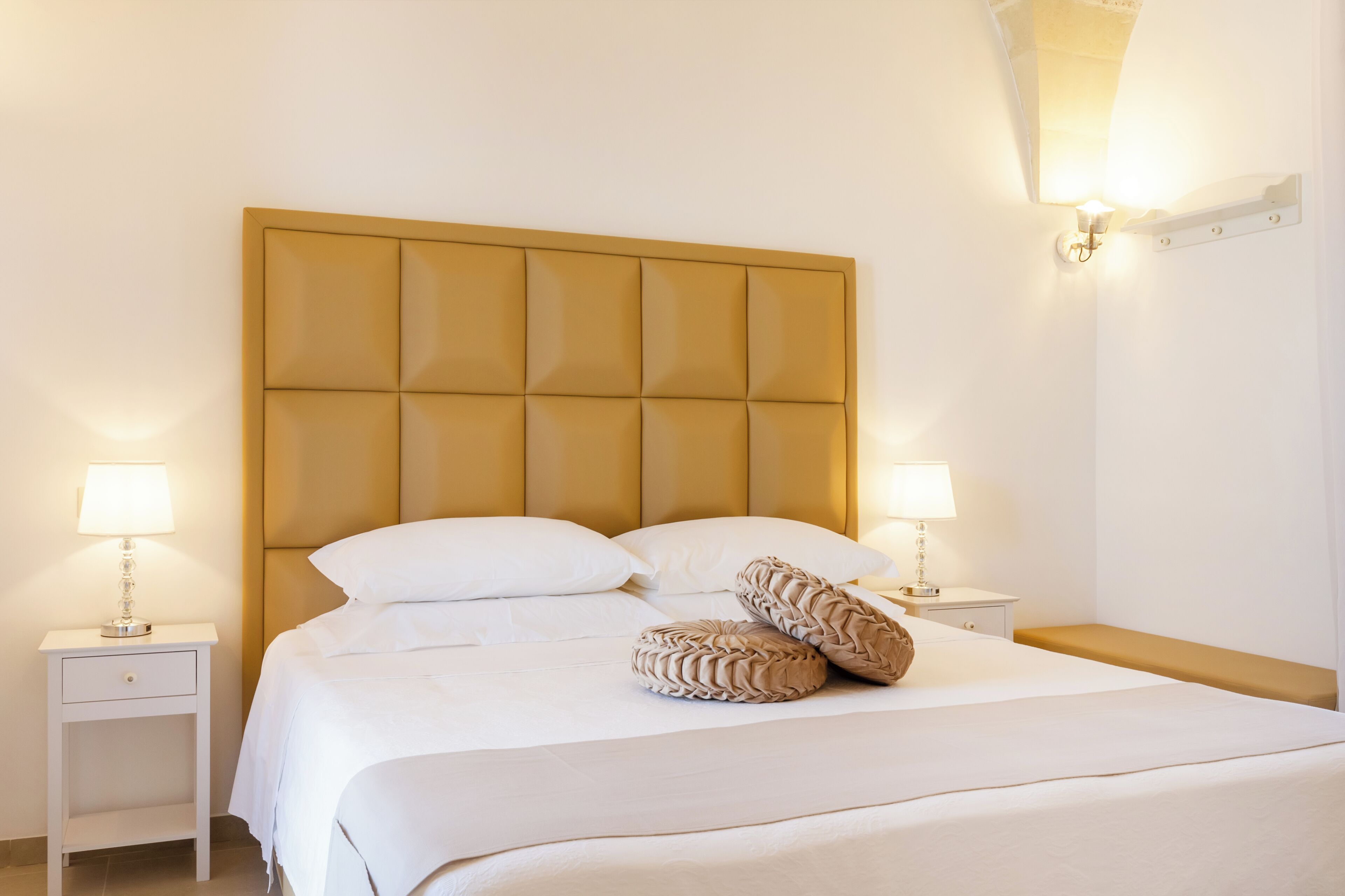 Una cama bien hecha con cabecera capitonada, dos mesitas de noche con lámparas y un banco al pie de la cama.